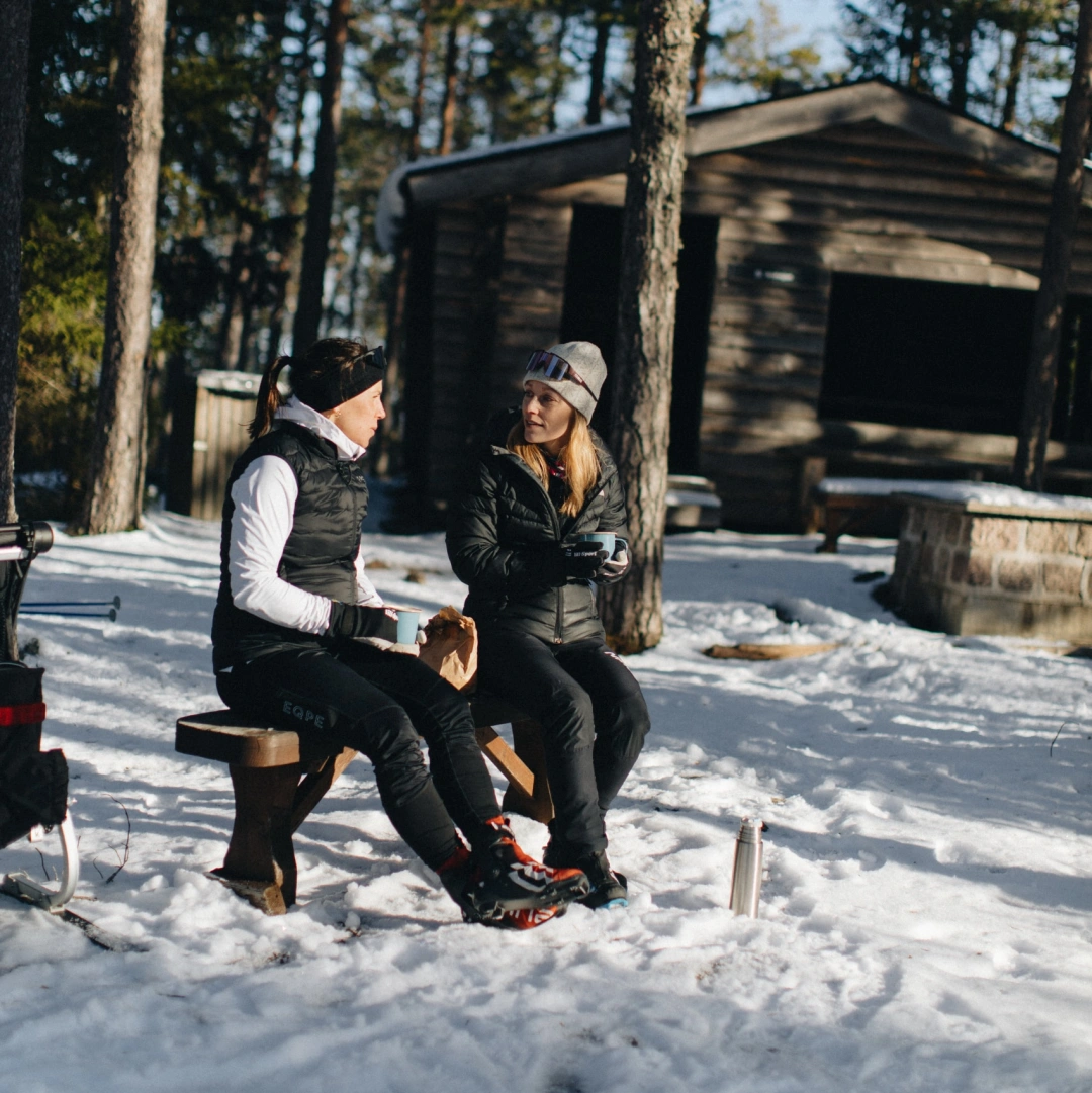 Två personer dricker kaffe från en termos på en bänk i snön